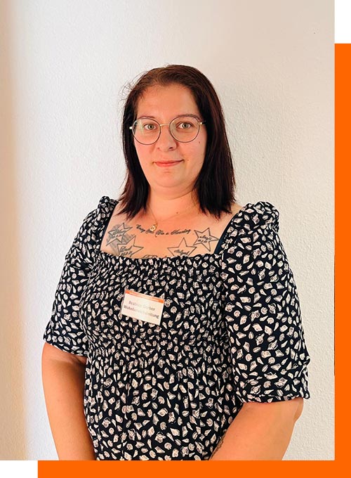 Person mit Brille und schwarzem Haar lächelt leicht; trägt gemustertes Kleid und hat Stern-Tattoos.