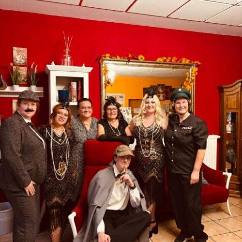 Sieben Personen in Kostümen der 1920er Jahre lächeln vor roter Wand und Spiegel