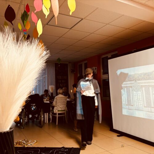 Eine stehende Person mit weißem Hemd und schwarzer Maske liest vor einer Leinwand im bunt geschmückten Raum