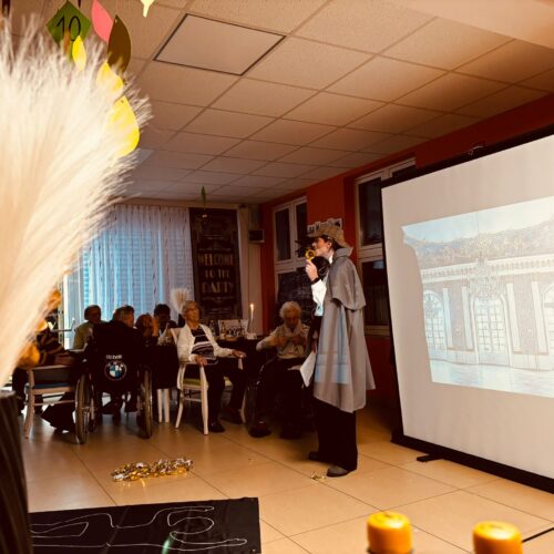 Ein älterer Mann mit Kostüm und Hut spricht lebhaft vor einer Gruppe am Beamer