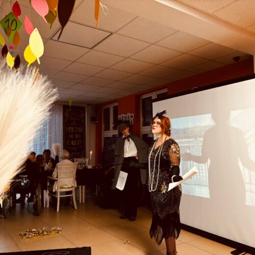 Eine Frau in festlicher, dunkler Kleidung mit Perlenkette lächelt vor einer Leinwand