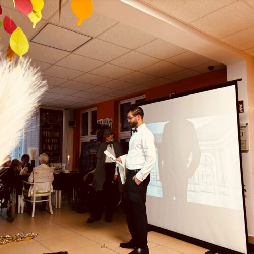 Ein Mann mit Bart hält lächelnd Notizen vor einer kleinen Gruppe in hellem Raum