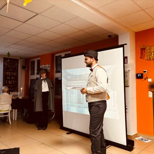 Ein Mann im Anzug mit Fliege und Zylinder steht vor einer Leinwand und spricht