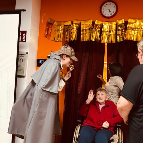 Eine Person im Detektivkostüm beugt sich lächelnd zu einer älteren Dame im Rollstuhl