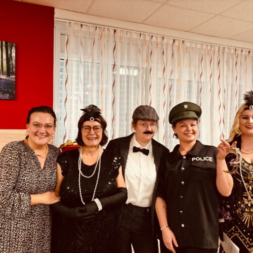 Fünf lachende Personen in bunten Kostümen stehen vor einem hellen Fenster zusammen