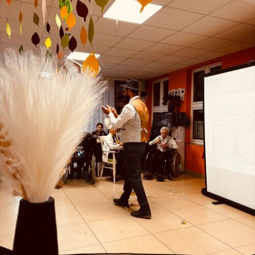 Ein Mann mit Hut und Weste spricht freundlich zu älteren Menschen im bunt dekorierten Raum