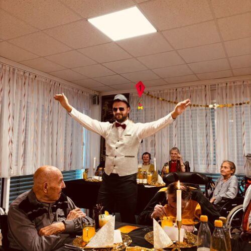 Ein Mann mit Fliege breitet lächelnd die Arme aus, während ältere Gäste am Tisch sitzen