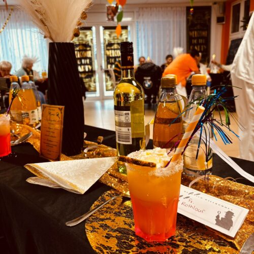 Ein festlich gedeckter Tisch mit goldenen Untersetzern, einer orangenen Cocktail und bunten Dekorationen vor weißen Vorhängen