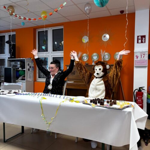 Zwei Personen, eine davon im Affenkostüm, heben fröhlich die Arme an einem bunt dekorierten Tisch