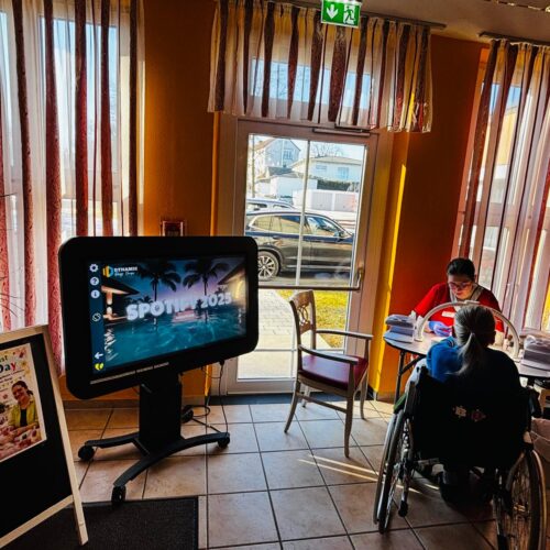 Zwei Personen, eine im Rollstuhl, sitzen freundlich an einem Tisch in einem sonnigen Café mit gelb-roten Wänden, daneben steht ein großer Bildschirm mit Palmenmotiv