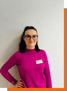 Eine lächelnde Frau mit Brille trägt einen pinken Pullover und Namensschild