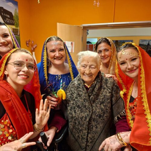 Fünf lächelnde Frauen in bunten traditionellen Kleidern vor einer orangefarbenen Wand